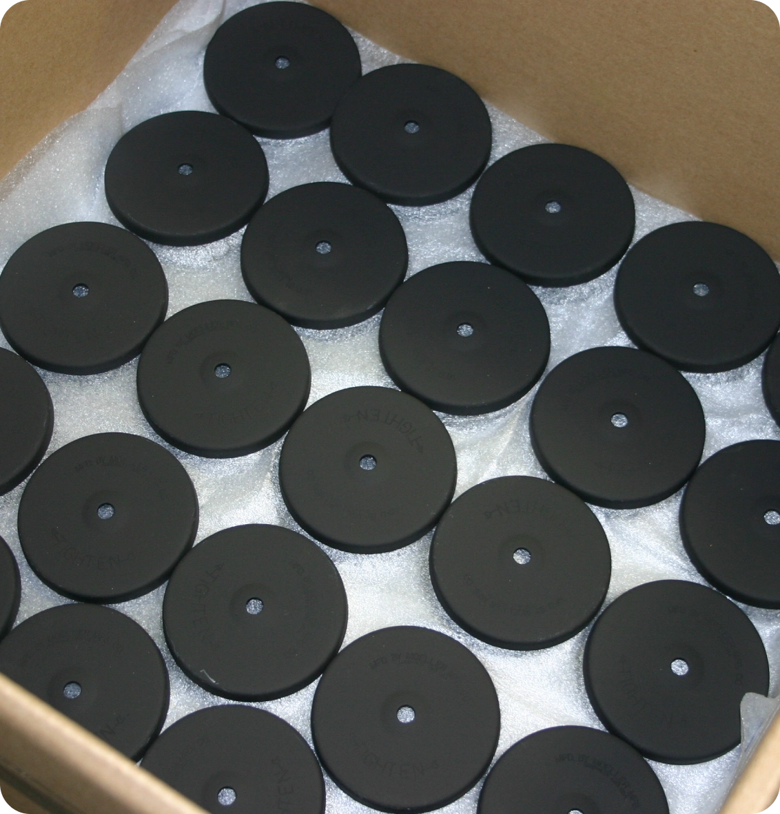 A cardboard box filled with rows of black hockey pucks on a layer of foam padding.