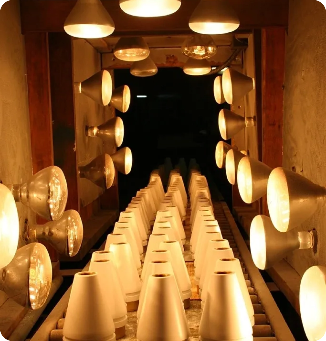 A conveyor belt with ceramic pots passes under rows of illuminated heat lamps inside an industrial kiln.